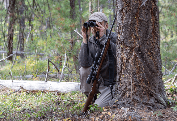 Hunter with binoculars behind tree in forest.